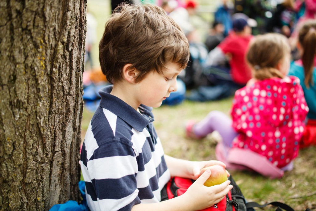 Young kid with bad social skill siting alone while other kids in the background.