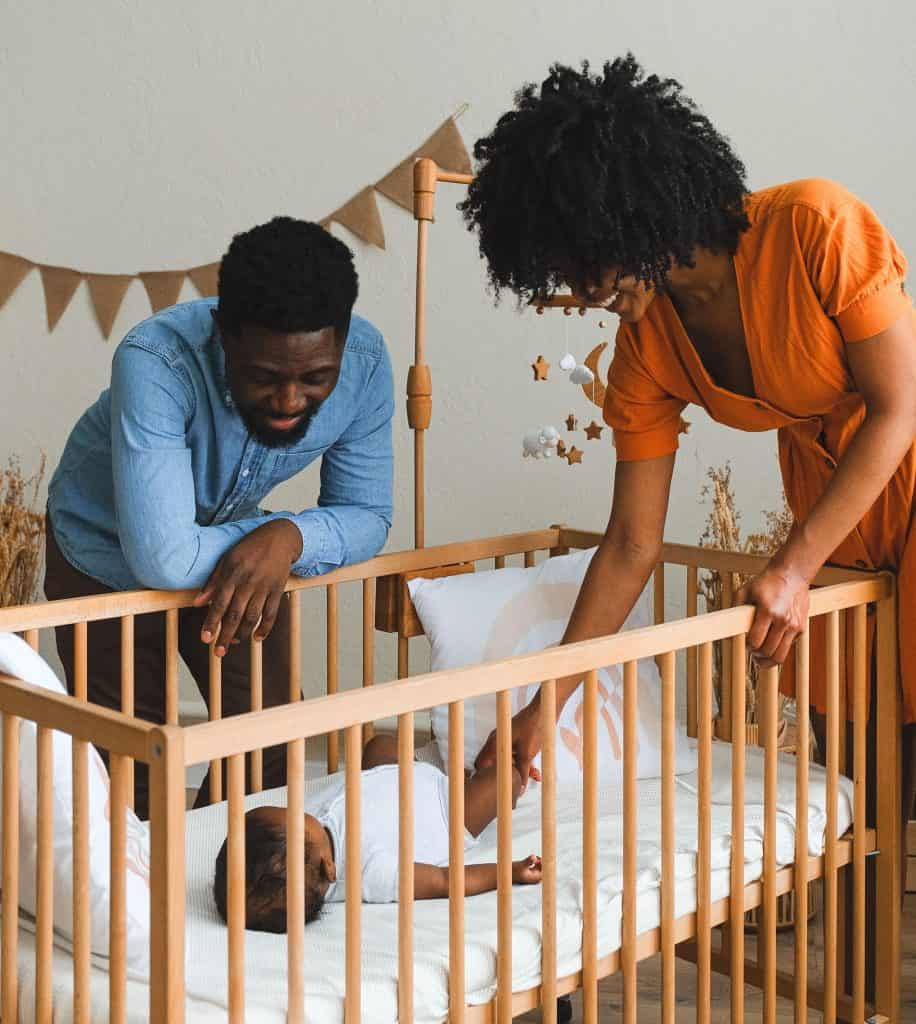 Two parents smiling looking down at toddler in a crib sleeping.