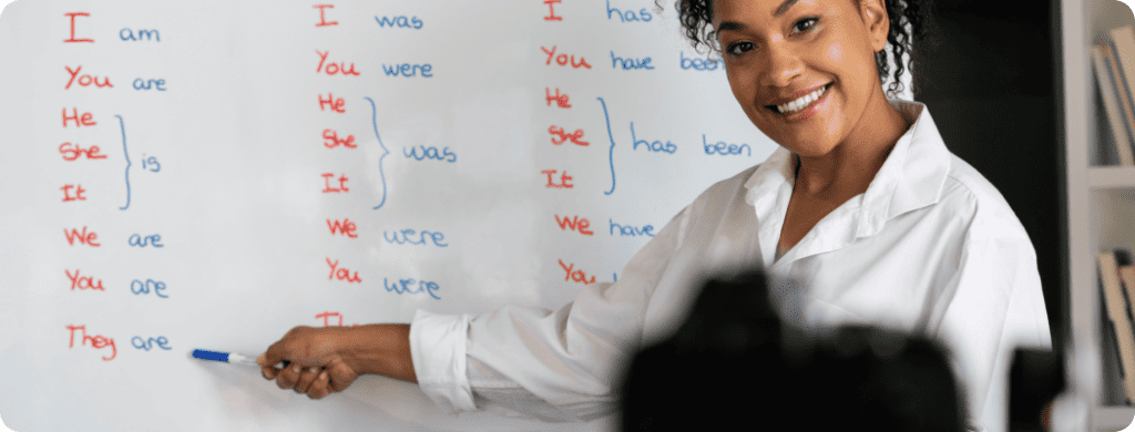 Tutor smiling while teaching and pointing at board in classroom.