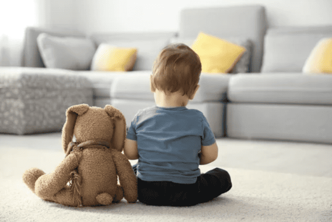 Sad toddler sitting on living room floor with teddy bear facing away.
