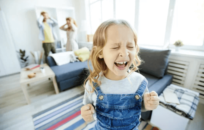 Child with Oppositional Defiant Disorder throwing tantrum out loud while parents are in the background covering their ears.